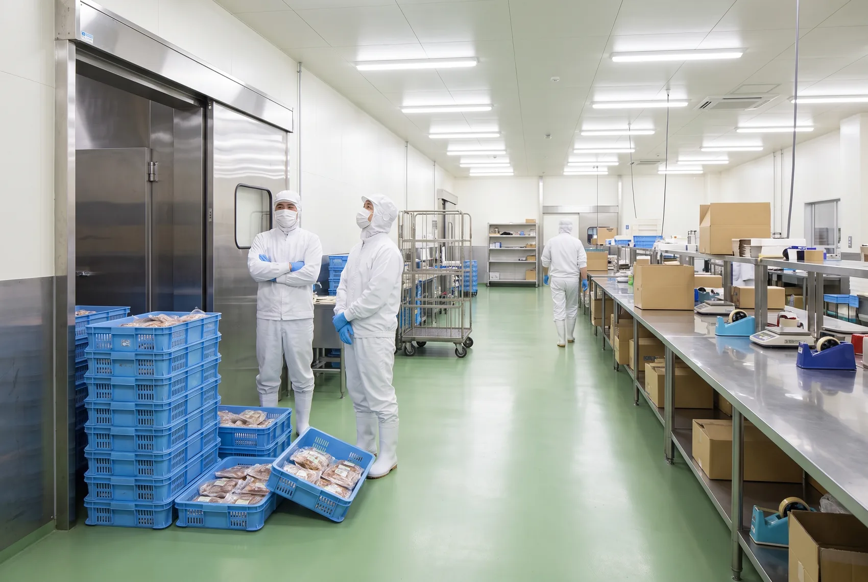 改善前。食品工場の受け渡し口に仕掛品のコンテナが高く積み上がり、床にも直置きされている。手前の2人は手を止めて山と扉を見ており、奥の1人は持ち場を離れて歩いている